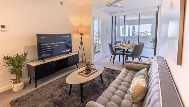 A modern living room with a flat-screen TV, a sofa, a coffee table, and a dining area with a glass door leading to a balcony. - Toowong Central, Carpark+Wifi, Great value 1BR Apt (Toowong)
