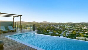 An infinity pool with a wooden deck and lounge chairs, overlooking a residential area with houses and hills. - Toowong Central, Carpark+Wifi, Great value 1BR Apt (Toowong)