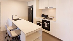 A modern kitchen with white cabinetry, a central island with bar stools, a built-in oven, and a stainless steel refrigerator. - Toowong Central, Carpark+Wifi, Great value 1BR Apt (Toowong)