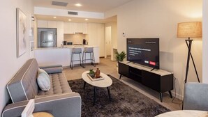 A modern living room with a sofa, coffee table, TV, and a small kitchen area in the background. - Toowong Central, Carpark+Wifi, Great value 1BR Apt (Toowong)