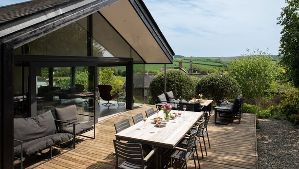 A modern outdoor dining area with a wooden deck, glass walls, and a view of the countryside. - Quarry House -  An architecturally inspiring house with sea views (West Charleton)