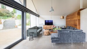 A modern living room with a large TV, grey sofas, and a wooden accent wall. - Quarry House -  An architecturally inspiring house with sea views (West Charleton)