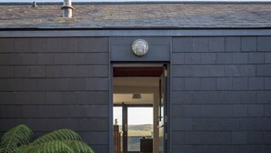 A modern building with a dark facade, a wooden pathway leading to an open doorway, and a view of a landscape through the doorway. - Quarry House -  An architecturally inspiring house with sea views (West Charleton)