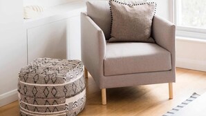 Ein modernes Wohnzimmer mit grauem Sessel, Hocker mit Muster, einem weißen Regal mit Büchern und Dekoration sowie einem großen Fenster, das natürliches Licht hereinlässt.