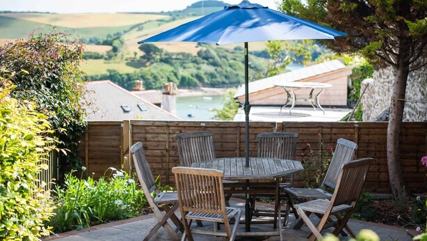 Eine Terrasse mit rundem Tisch und vier Stühlen, einem blauen Sonnenschirm und Blick auf einen See und Hügel im Hintergrund.