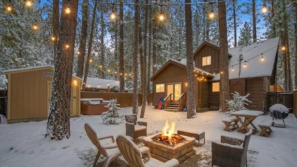 Eine Hütte mit Feuerstelle, Sitzgelegenheiten im Freien und Lichterketten in verschneiter Waldlandschaft.