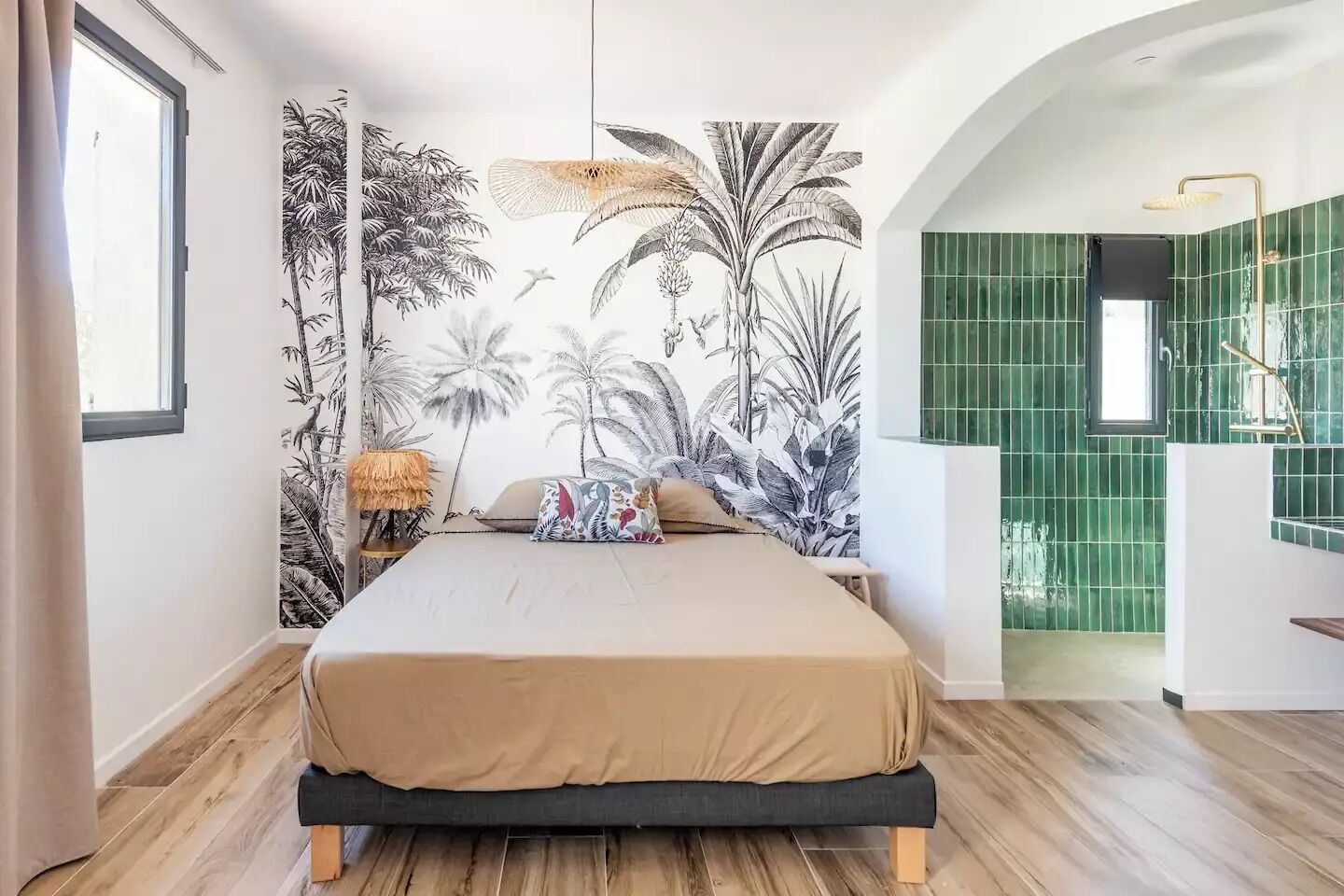 Une chambre avec un grand lit, une fresque représentant des plantes tropicales, une petite table avec une lampe, et une salle de bain aux carreaux verts visible à travers une arche.