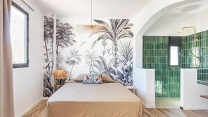 Un dormitorio con una cama grande, un mural de plantas tropicales, una mesita de noche con una lámpara, y un baño con azulejos verdes visible a través de un arco.
