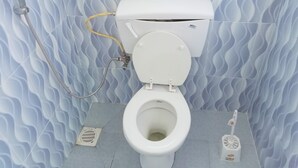 A toilet with a white seat and lid, a yellow hose, and a white bidet attachment on the floor. - Naivasha Homestay (Naivasha)
