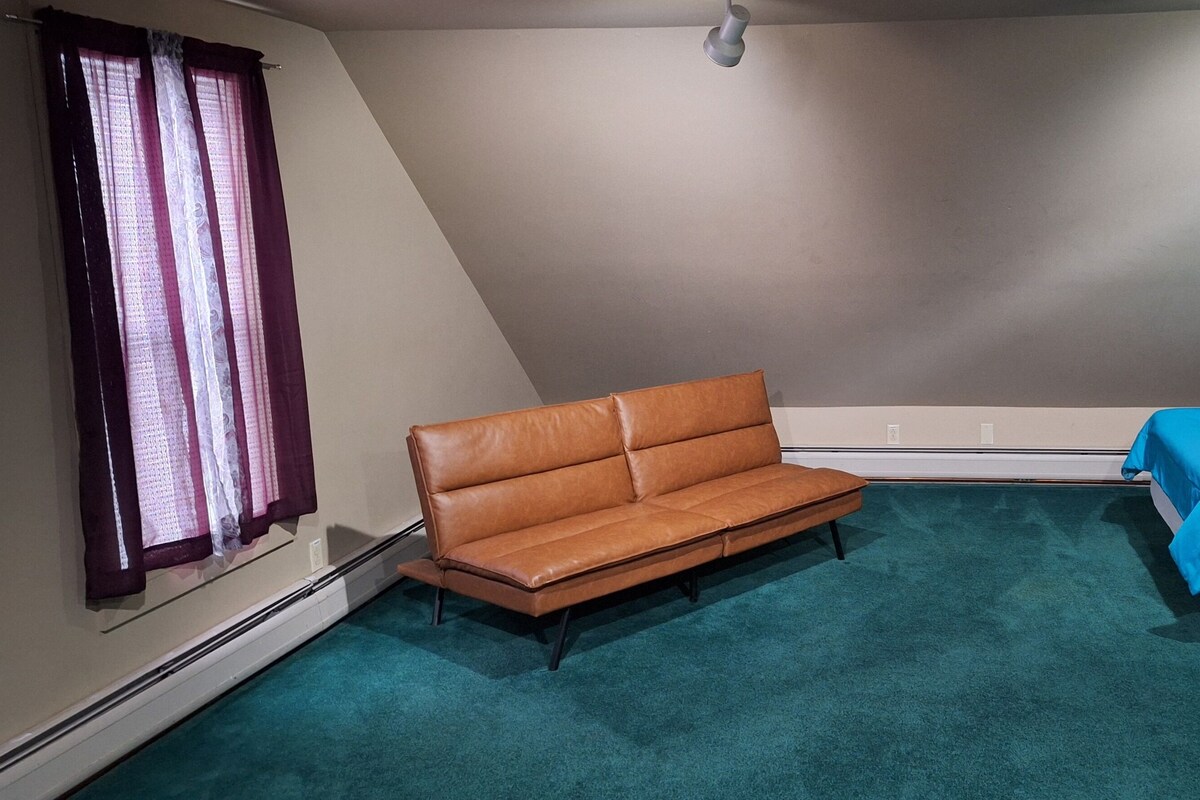A small, triangular room with a brown leather sofa, a bed with a blue bedspread, and a window with purple curtains.