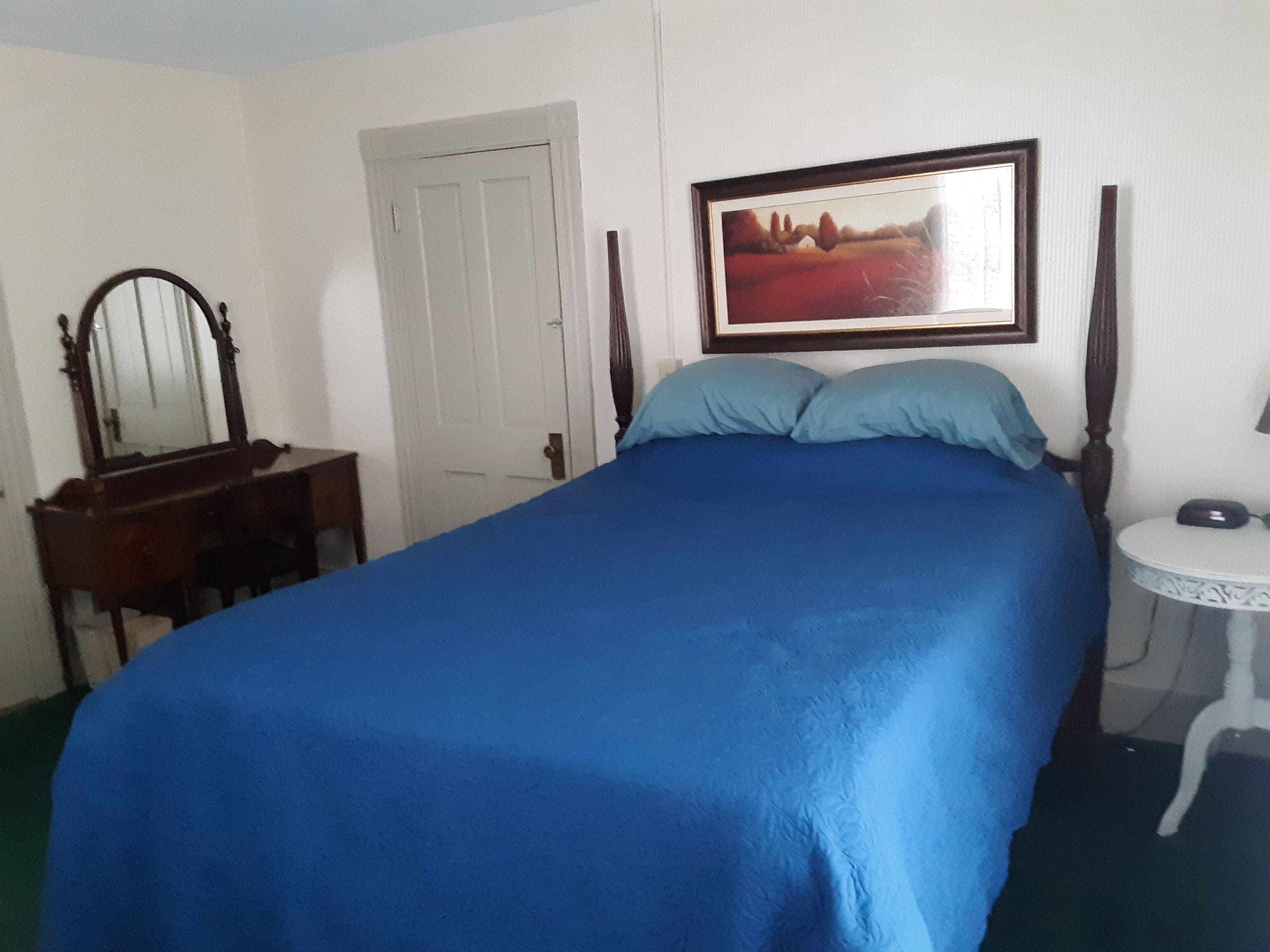 Una habitación con una cama, un tocador, un espejo, una puerta y un cuadro en la pared.