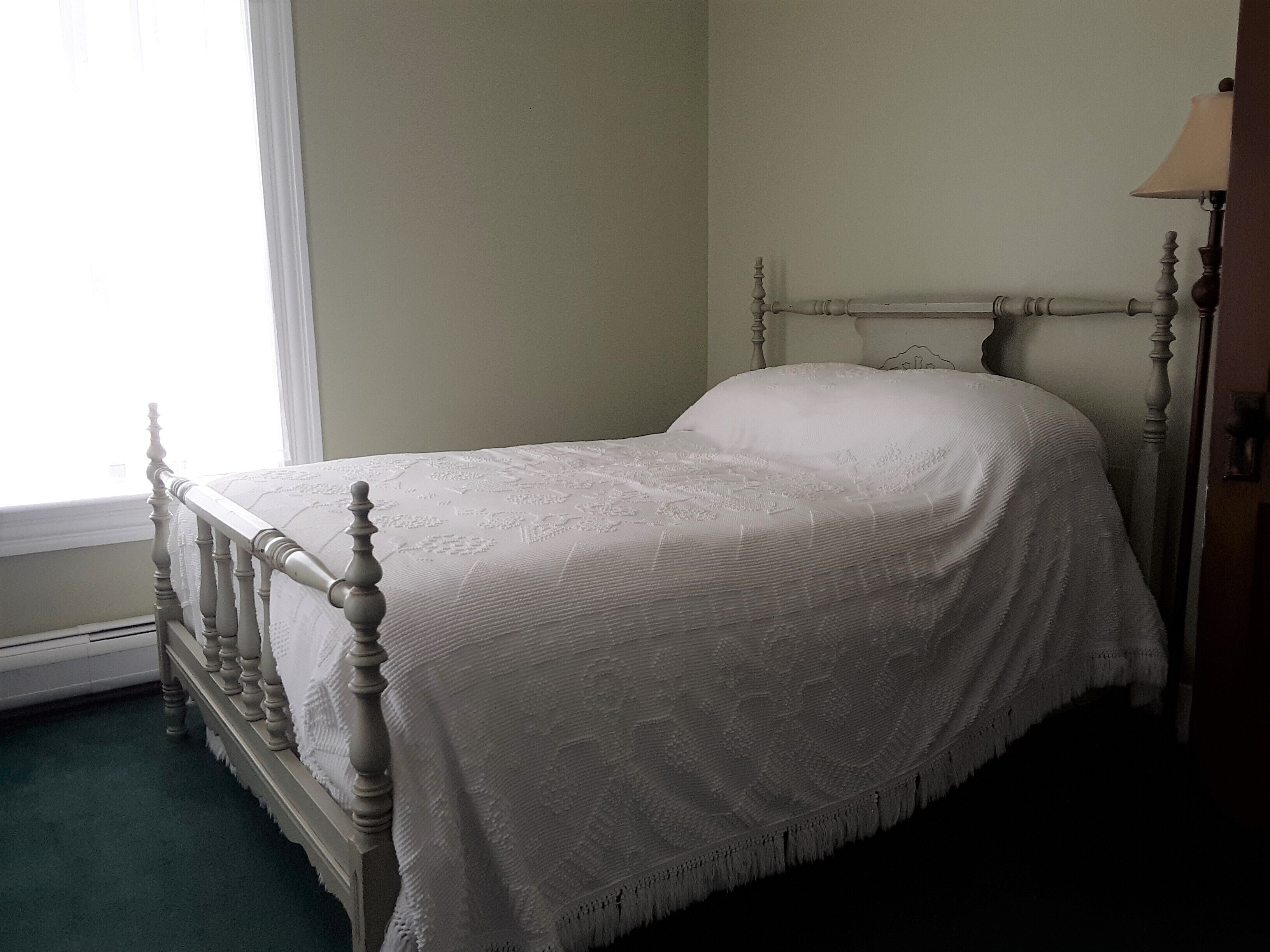 A bed with a wooden frame and a white bedspread in a room with a window and a lamp.