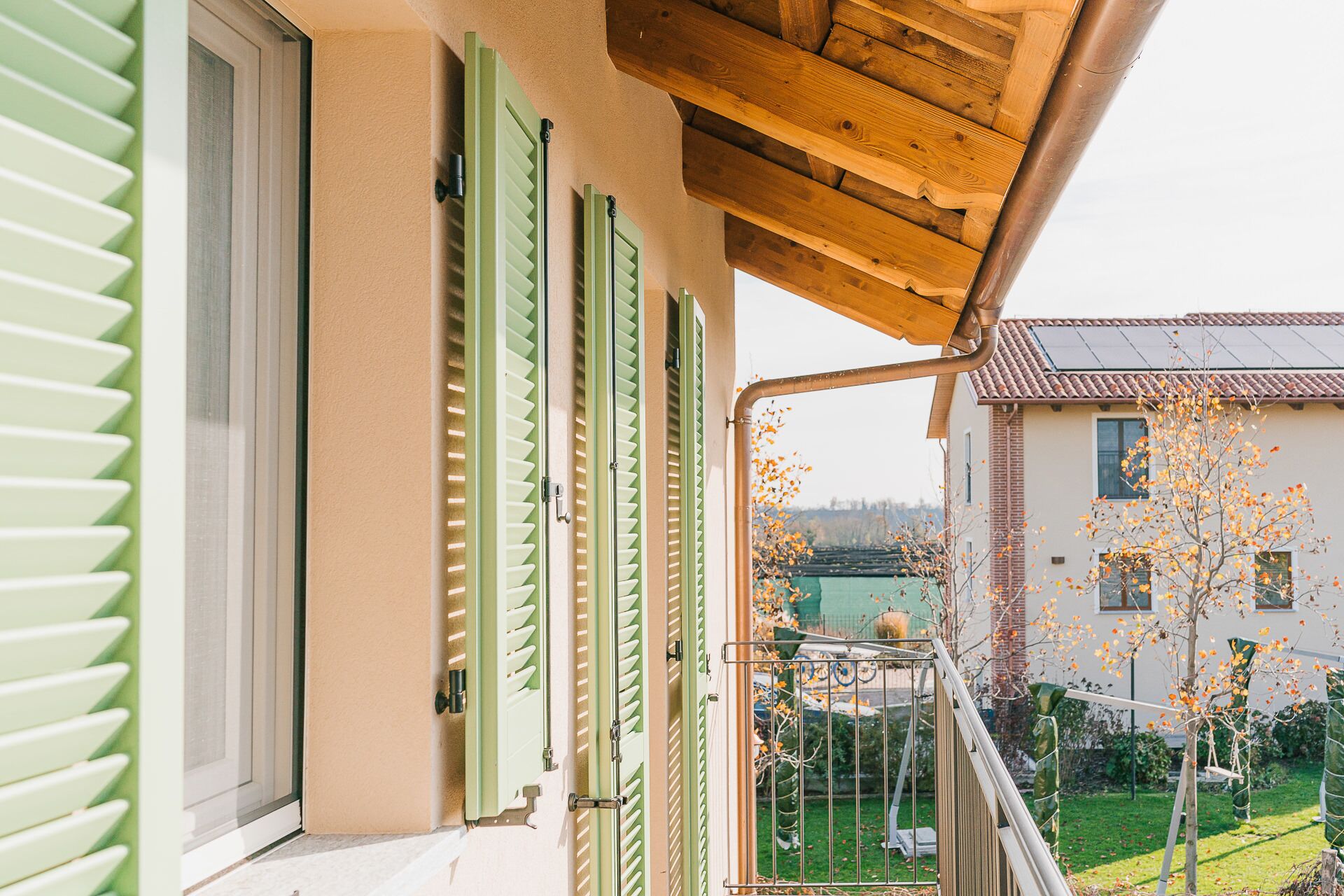 Una casa con persiane verdi, un balcone in legno e vista sulle case e gli alberi vicini.