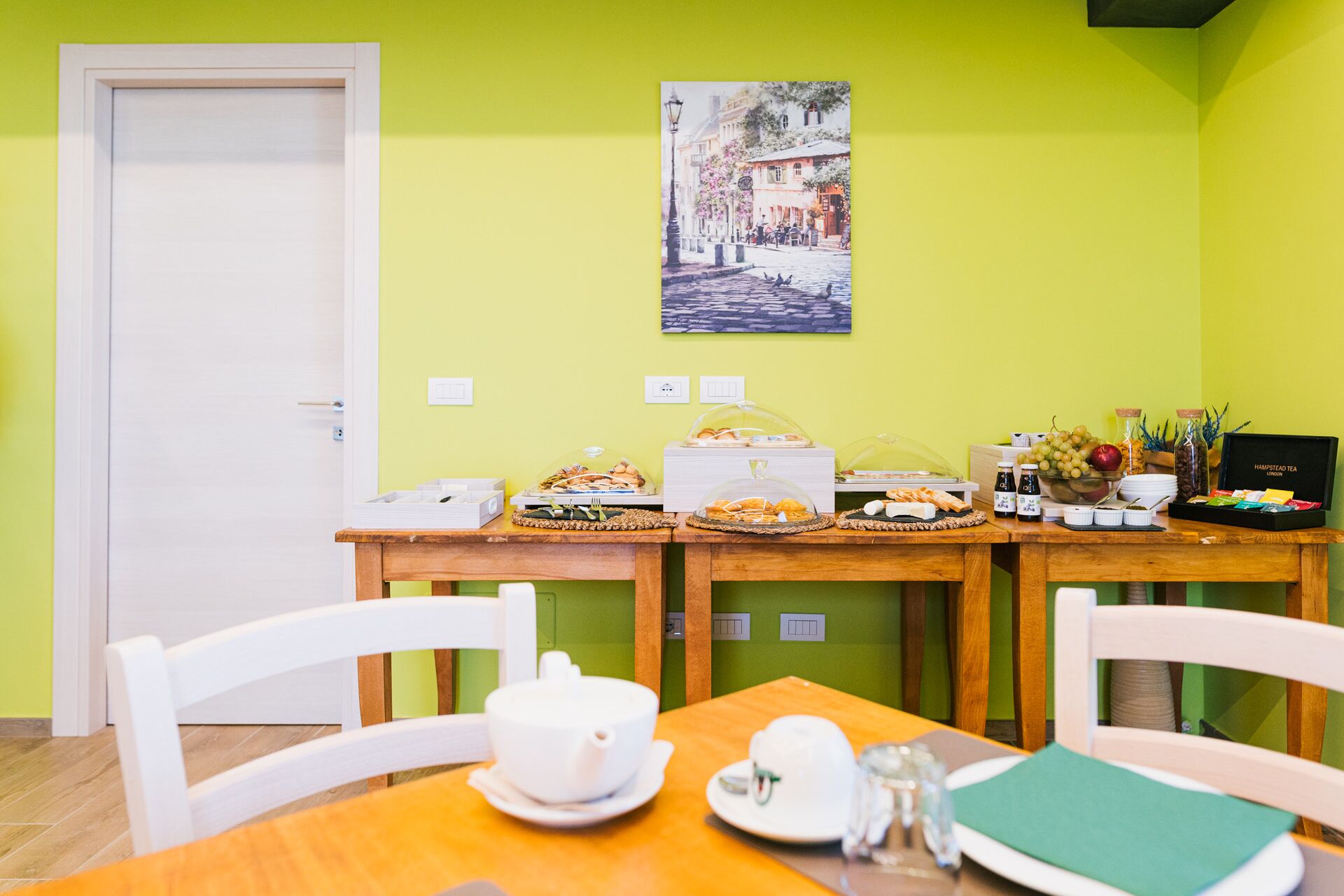 Um buffet de café da manhã com diversos alimentos e uma caixa de chá sobre uma mesa de madeira em frente a uma parede verde.