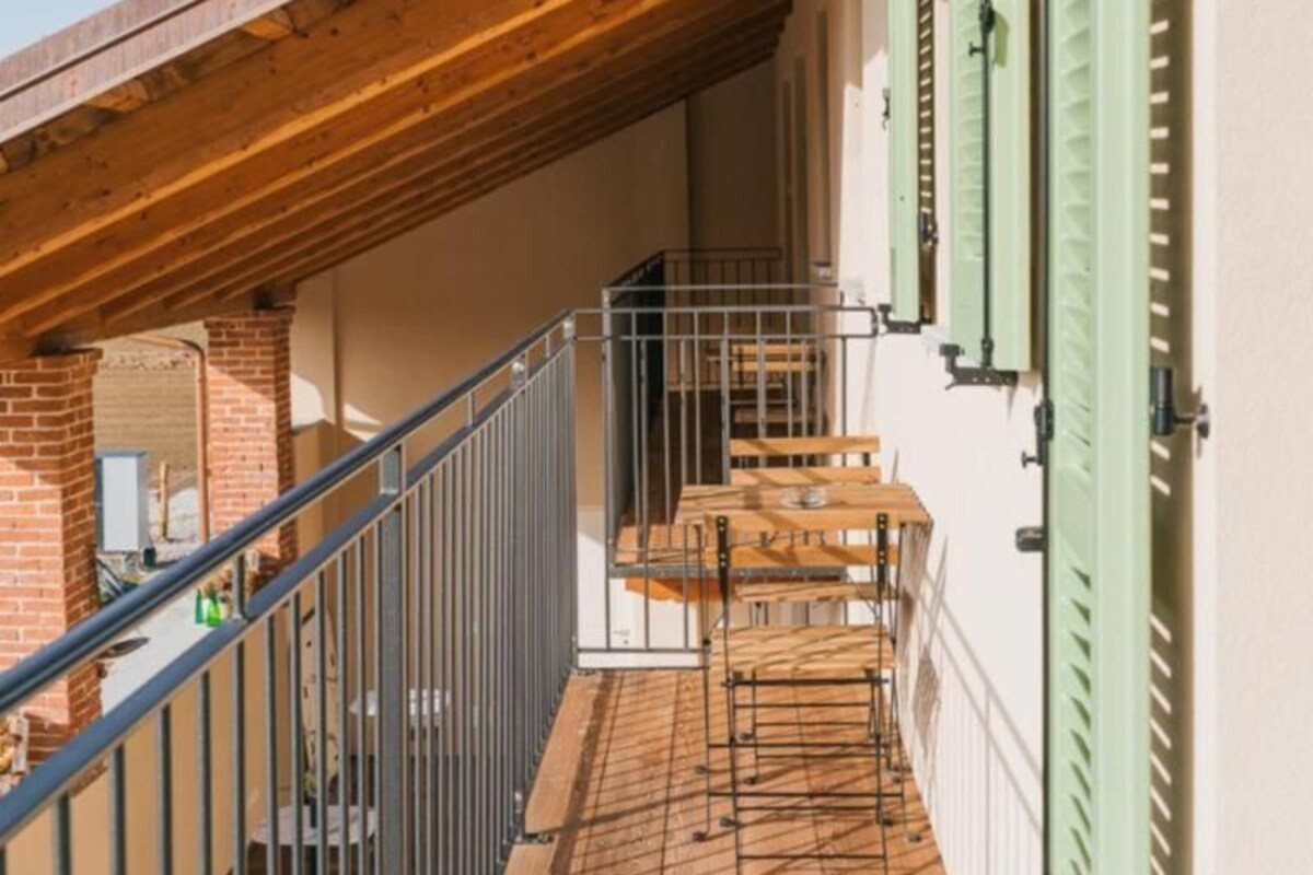 Ein hölzerner Balkon mit Metallgeländer, eine Treppe zu einer oberen Ebene und grüne Fensterläden am Gebäude.