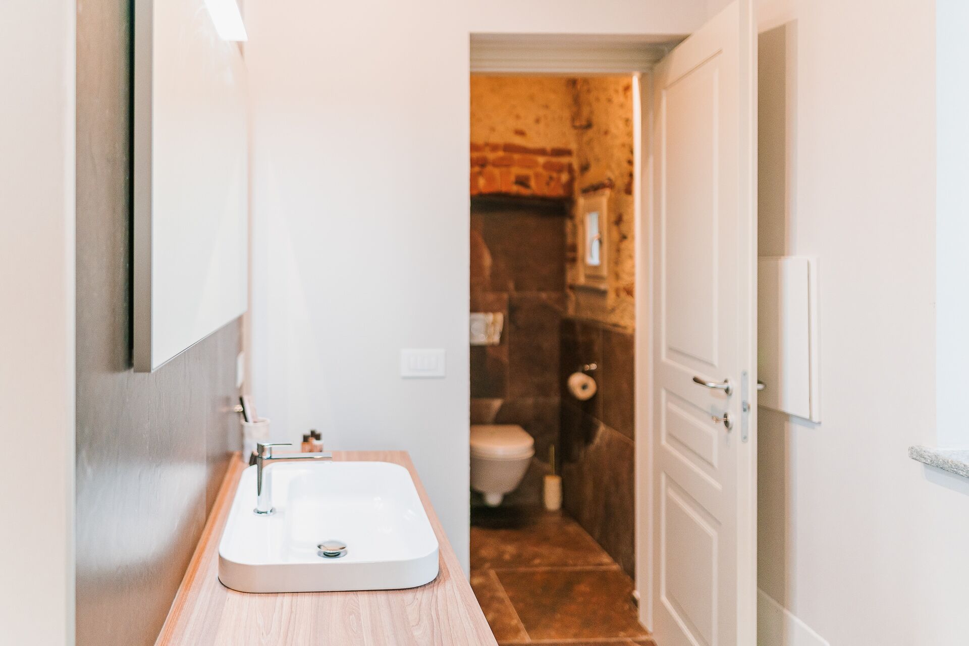 Une petite salle de bain avec un lavabo blanc, un plan de travail en bois et une porte donnant sur une pièce dont un mur est en pierre et qui contient des toilettes.