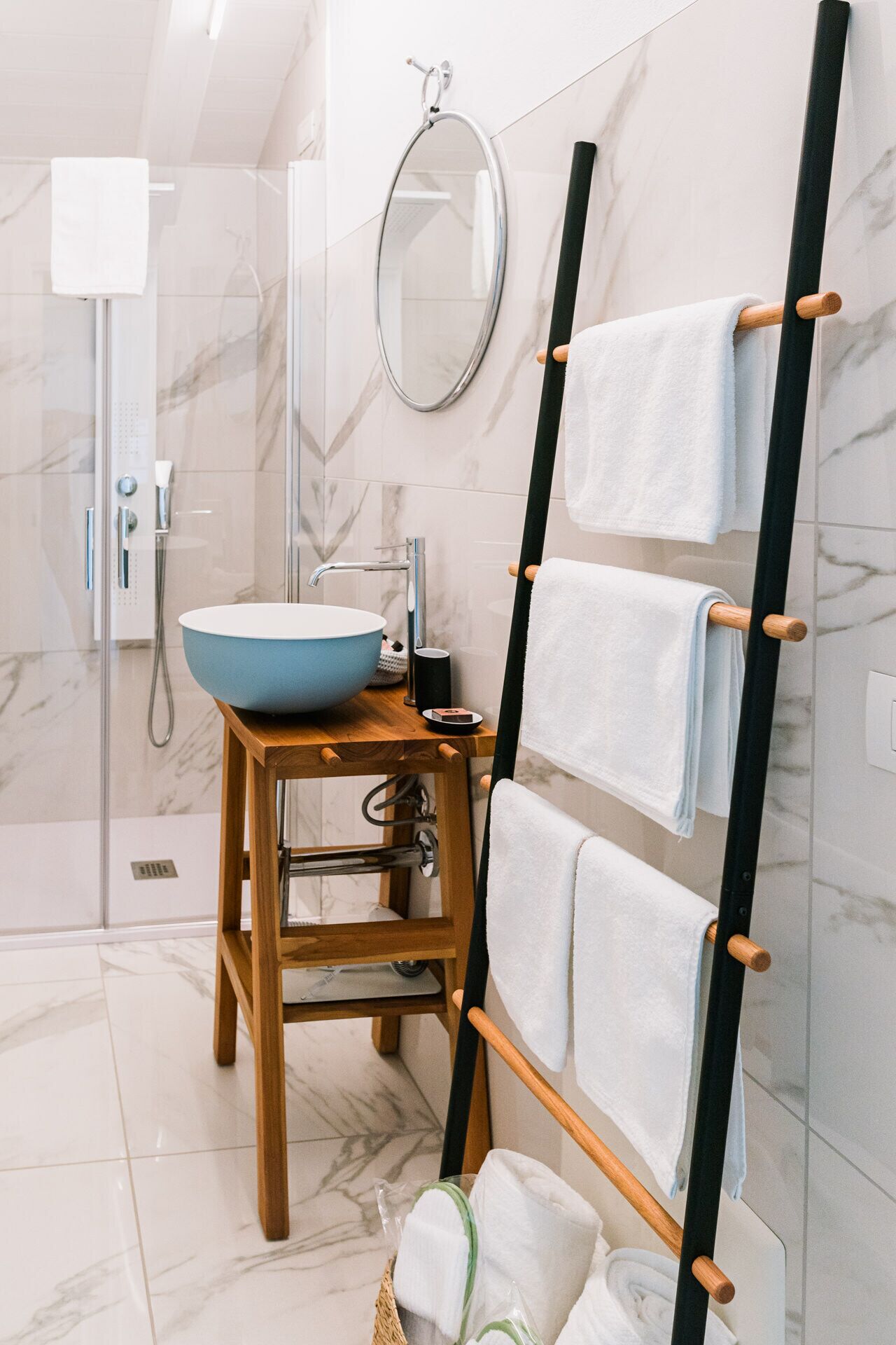 A modern bathroom with a wooden stool, a blue basin, a towel rack, and a shower area.