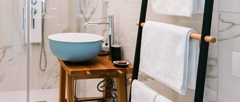 A modern bathroom with a wooden stool, a blue basin, a towel rack, and a shower area.