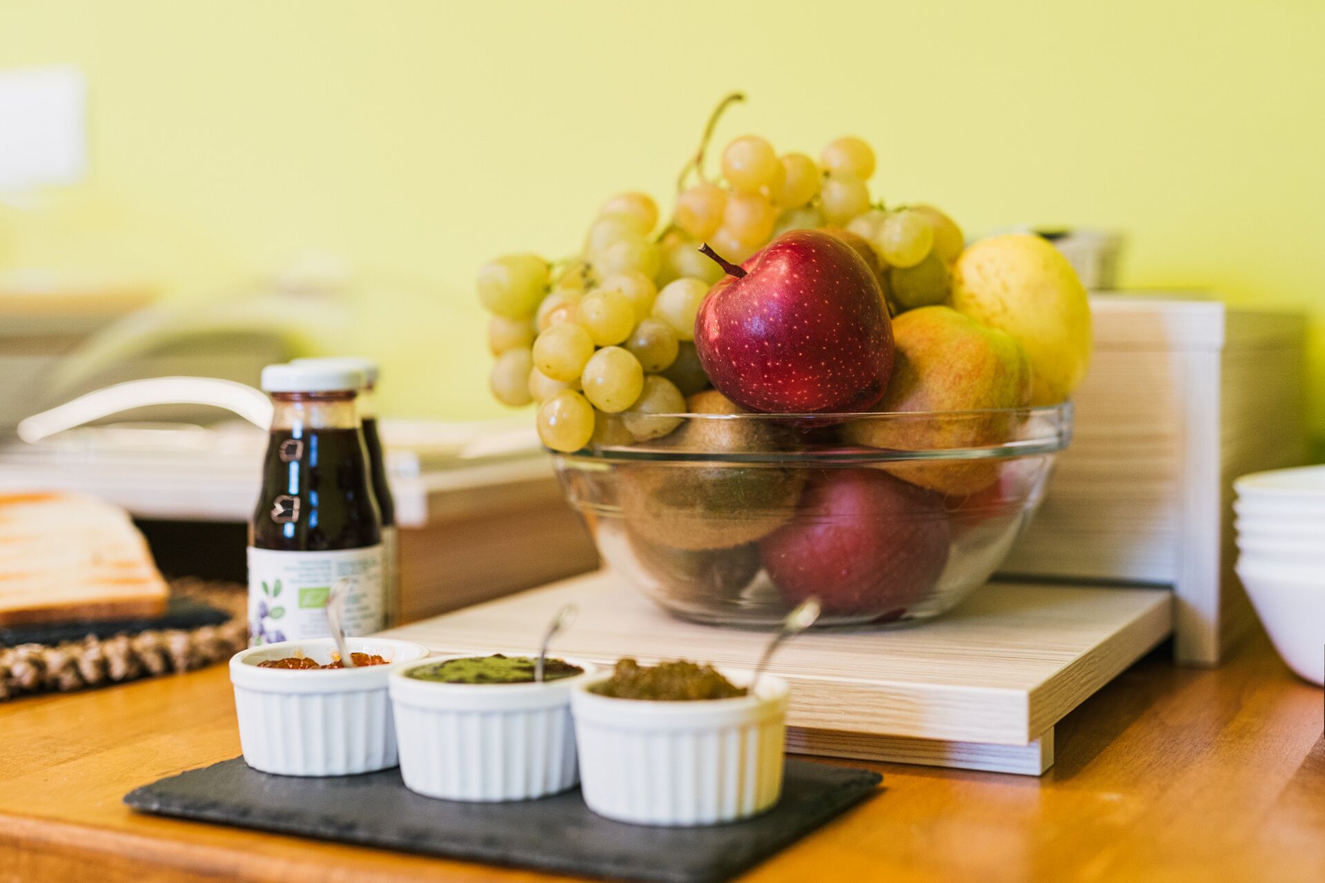 Un saladier rempli de fruits, notamment des raisins, des pommes et des poires, avec de petits récipients contenant des condiments, le tout posé sur une table en bois.
