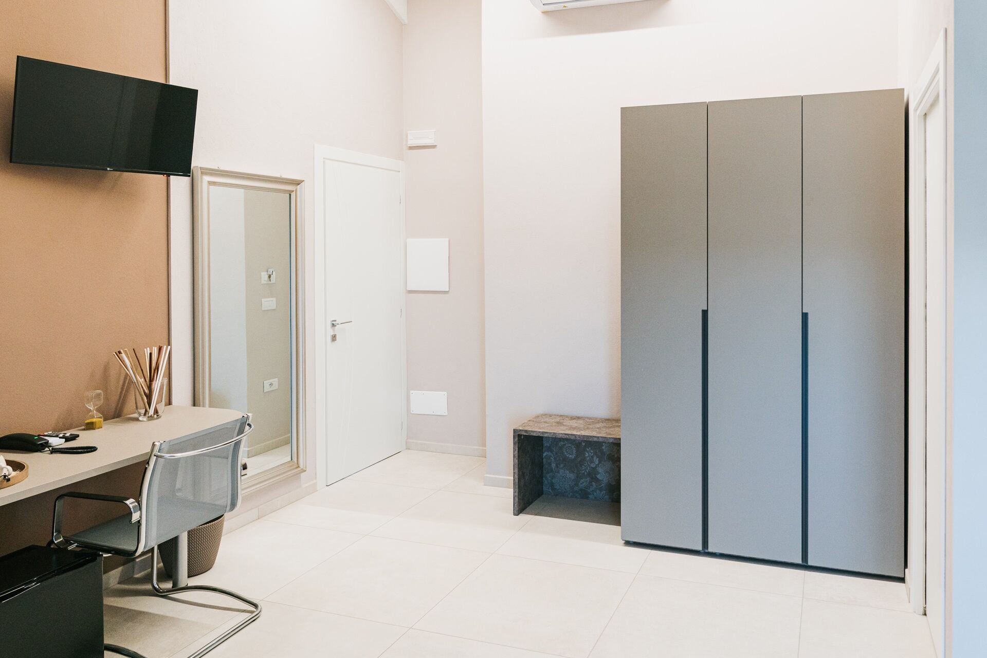 A modern hotel room with a desk, chair, wardrobe, and a television mounted on the wall.