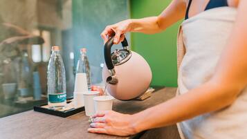 Una persona sirve agua de una tetera en una taza sobre una encimera con varios objetos, como botellas y tazas.