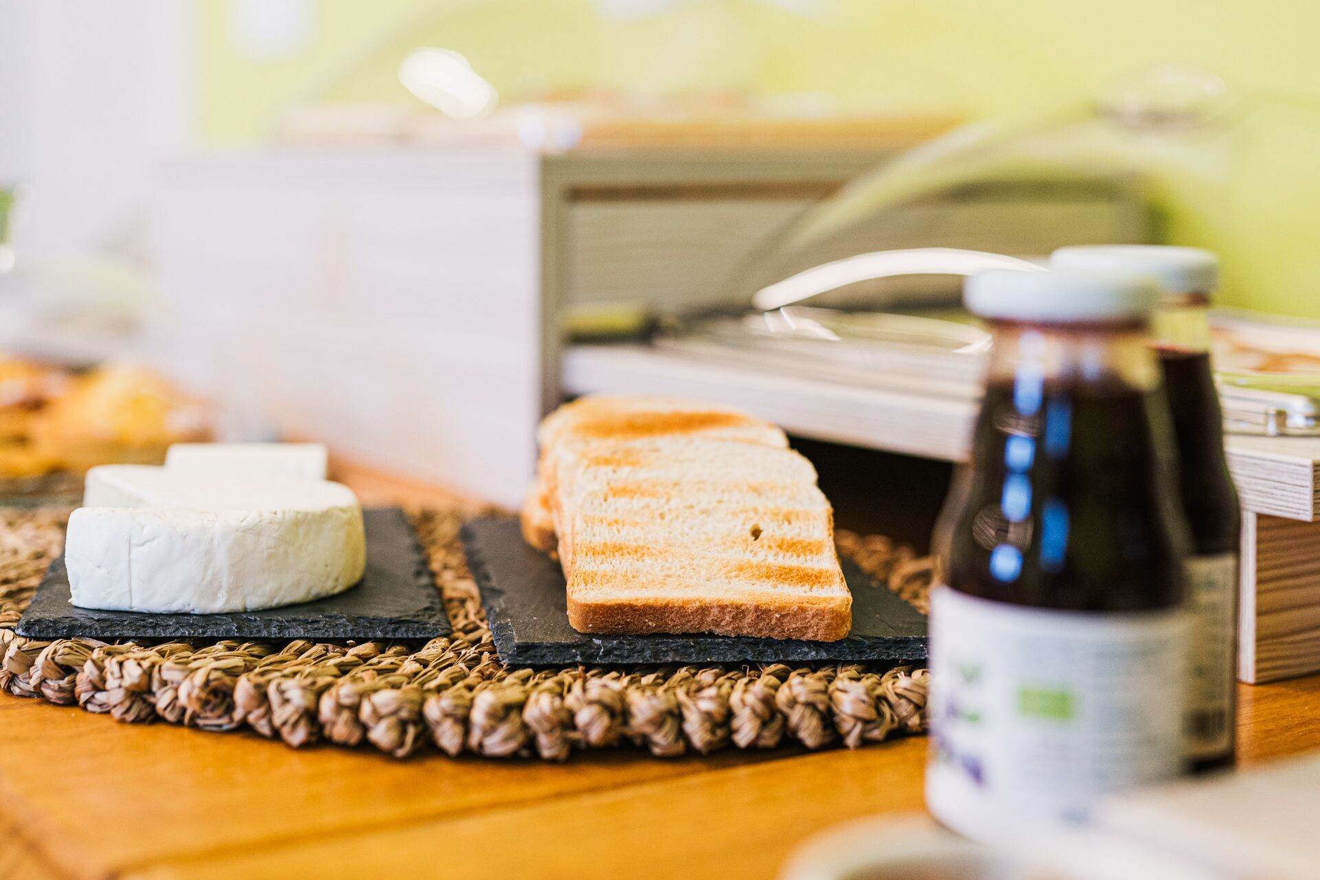 A cheese board with various cheeses and bread on a woven placemat, a bottle of sauce, and a coffee machine in the background.