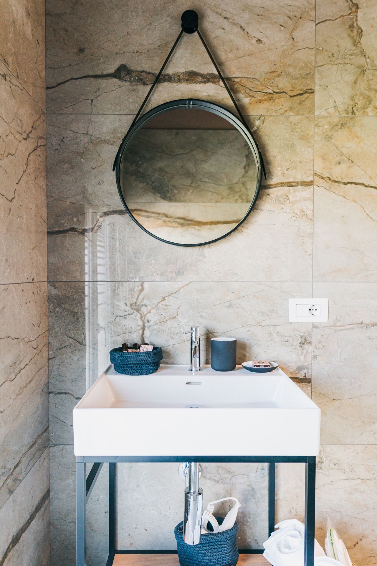 A bathroom with a marble wall, a round mirror, a white sink, a blue basket, and a blue vase.