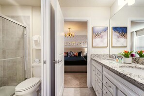 A bathroom with a shower, toilet, and sink. There is a bed with a headboard and pillows in the background. - Luxurious 8BR Villa Retreat Themed Rooms & Pool (Davenport)