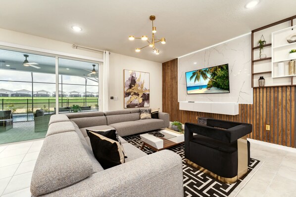A modern living room with a sectional sofa, a black and gold entertainment unit, a mounted TV, and a large window overlooking a landscaped area. - Luxurious 8BR Villa Retreat Themed Rooms & Pool (Davenport)