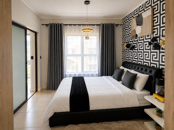 A modern bedroom with a large bed, a black headboard, a geometric-patterned wall, and a sliding glass door leading to a balcony.