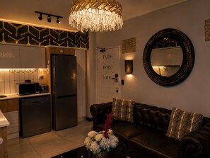 A modern kitchen with a refrigerator, microwave, and toaster, a dark leather sofa with decorative pillows, and a chandelier.