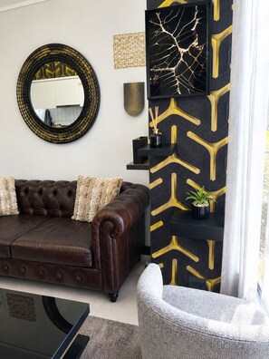 Una sala de estar con un sofá de cuero marrón, un espejo redondo y un zócalo con estampado negro y dorado.