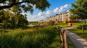 A residential complex with a pathway leading to a gated entrance, surrounded by greenery and a body of water.