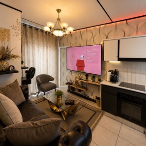 A modern living room with a sofa, armchair, and coffee table. A flat-screen TV is mounted on the wall, and a kitchen area is visible to the right.