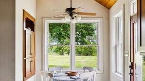 Comedor con mesa lista para dos personas, ventilador de techo y vistas a un paisaje verde a través de amplios ventanales.