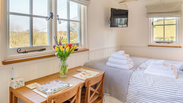 Una habitación con una mesa de madera, un jarrón con flores, una cama con una colcha a rayas y una ventana que da a la campiña.