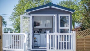 A small, single-story structure with a white exterior and a dark roof, featuring a kitchen area with cabinets and appliances visible through the open door. - The Studio - 1 Bedroom Cabin - Amroth (Narberth)