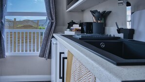 A modern kitchen with a black countertop, a built-in sink, and a window with curtains. - The Studio - 1 Bedroom Cabin - Amroth (Narberth)