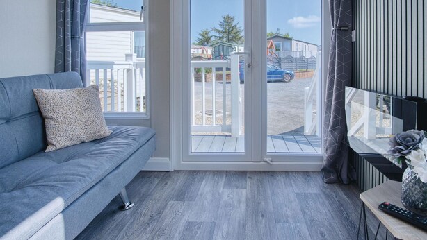 A modern living room with a grey sofa, a sliding glass door leading to an outdoor area, and a small table with a vase and remote control. - The Studio - 1 Bedroom Cabin - Amroth (Narberth)