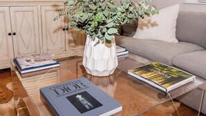 A living room with a clear glass coffee table, a white vase with green foliage, a sofa, and books. - Historic Charm | Near USC, Fort Jackson & 5 Points (Columbia)