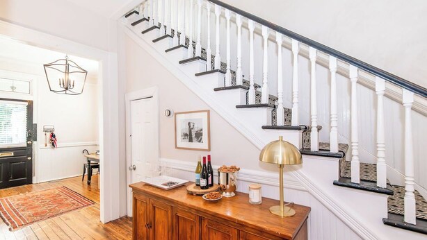 A staircase with white balusters and dark handrails, a wooden cabinet with a brass lamp, and a dining area with a red rug. - Historic Charm | Near USC, Fort Jackson & 5 Points (Columbia)
