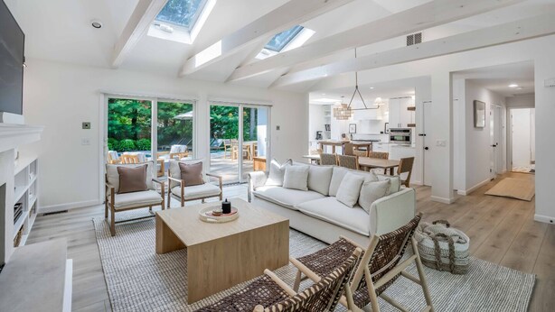 A spacious living area with a sofa, chairs, and a coffee table. There is a kitchen in the background and a view of greenery outside.