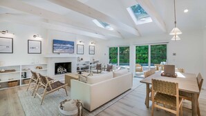 A spacious living area with a fireplace, a dining table, and a sofa. There are skylights on the ceiling, and the room has large windows.