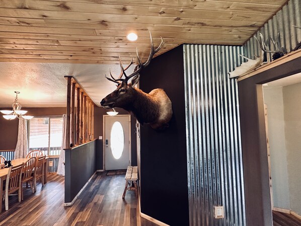 Una stanza con una testa di cervo appesa al muro, pavimento in legno, un'area pranzo con sedie e tavolo, e un soffitto con travi in legno.