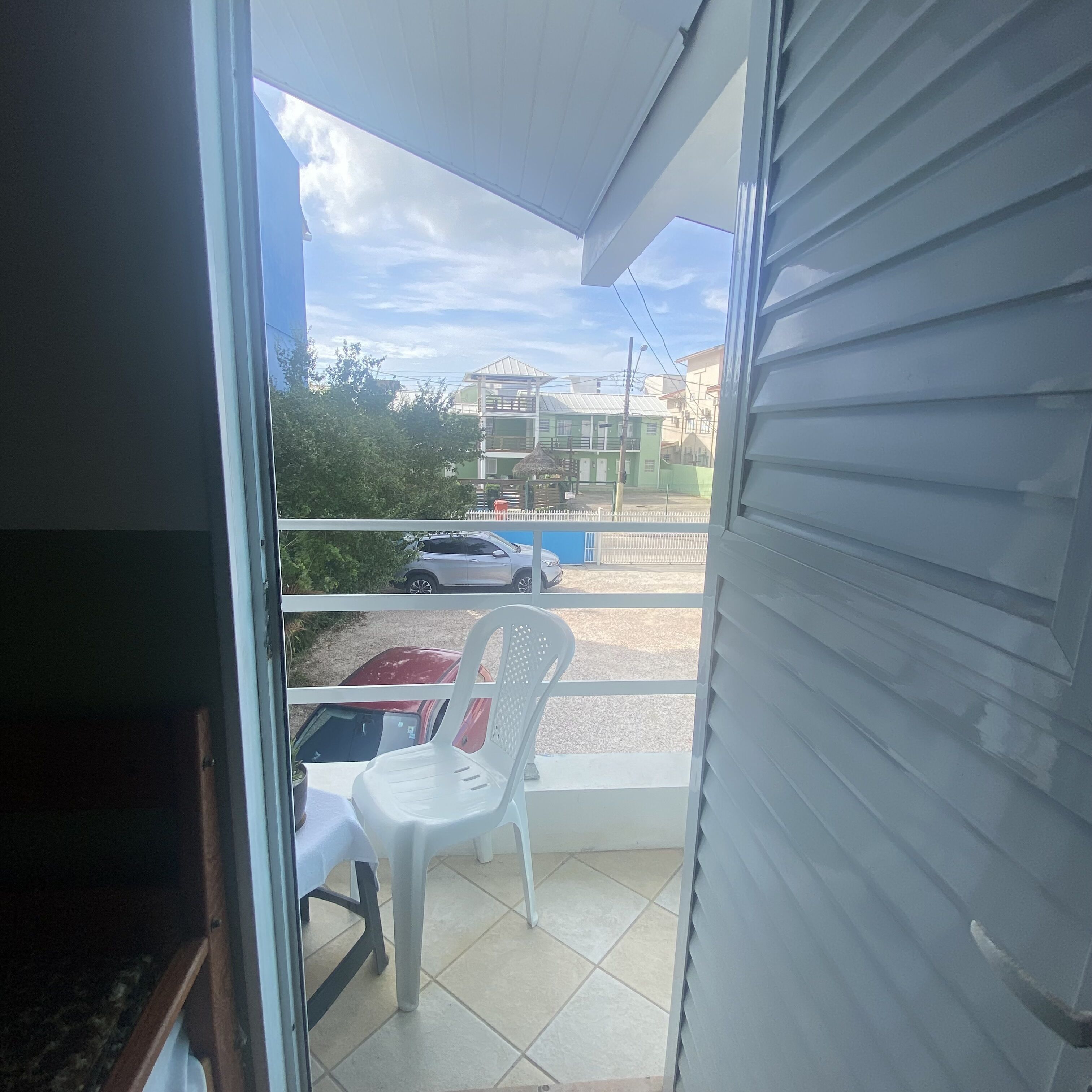 Ein Balkon mit einem weißen Plastikstuhl, einem Tisch mit weißer Tischdecke und Blick auf einen Parkplatz mit Autos und Gebäuden.