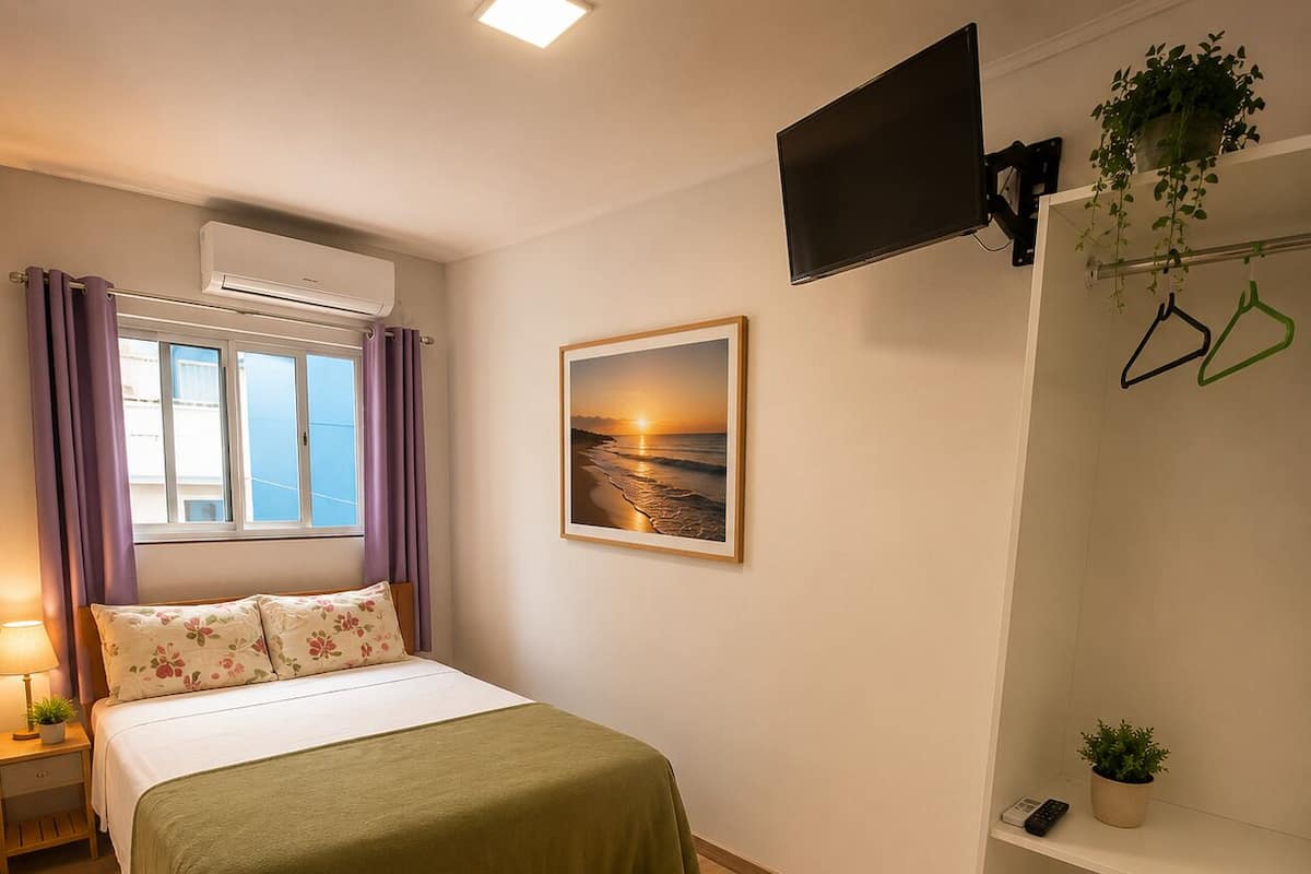 A bedroom with a bed, a window with curtains, a wall-mounted TV, a shelf with a plant, and a framed picture.