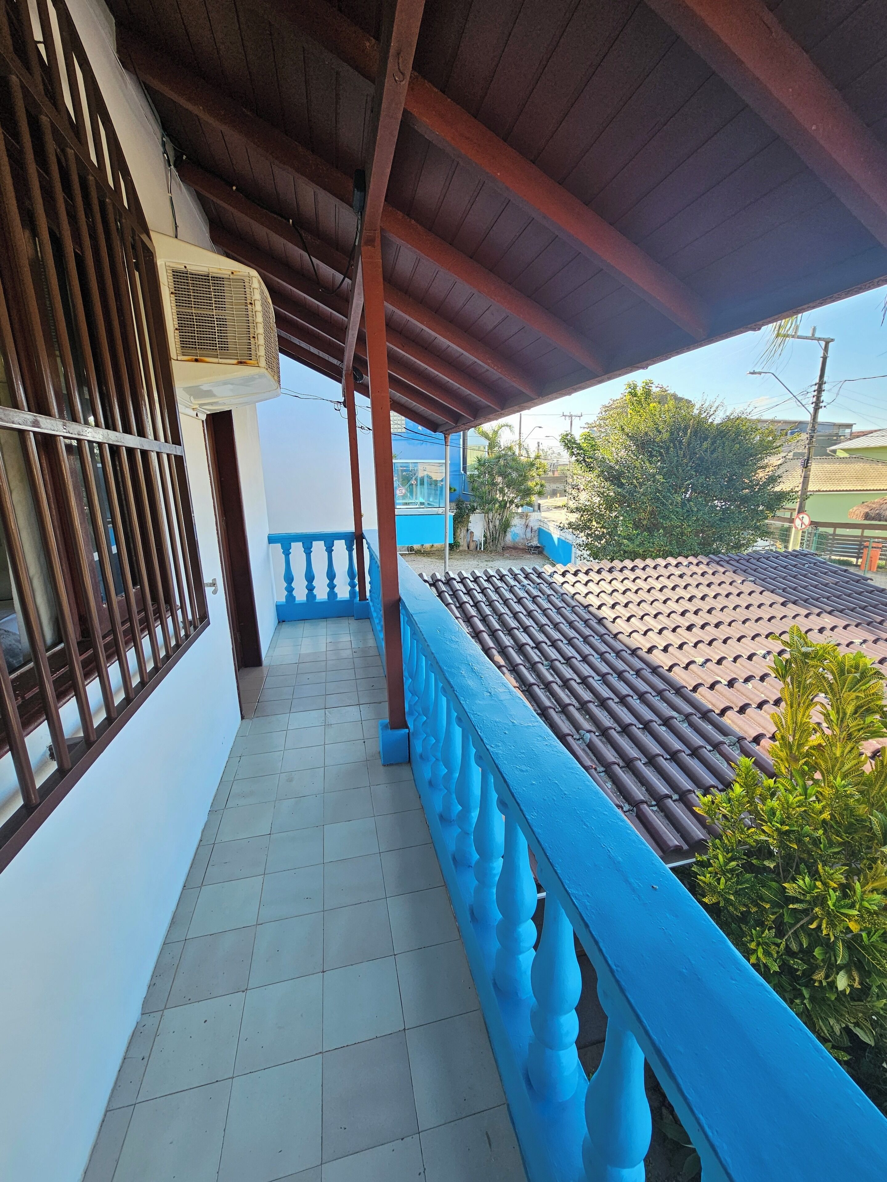 A covered balcony with a tiled floor, a blue railing, and a view of a residential area with houses and trees.