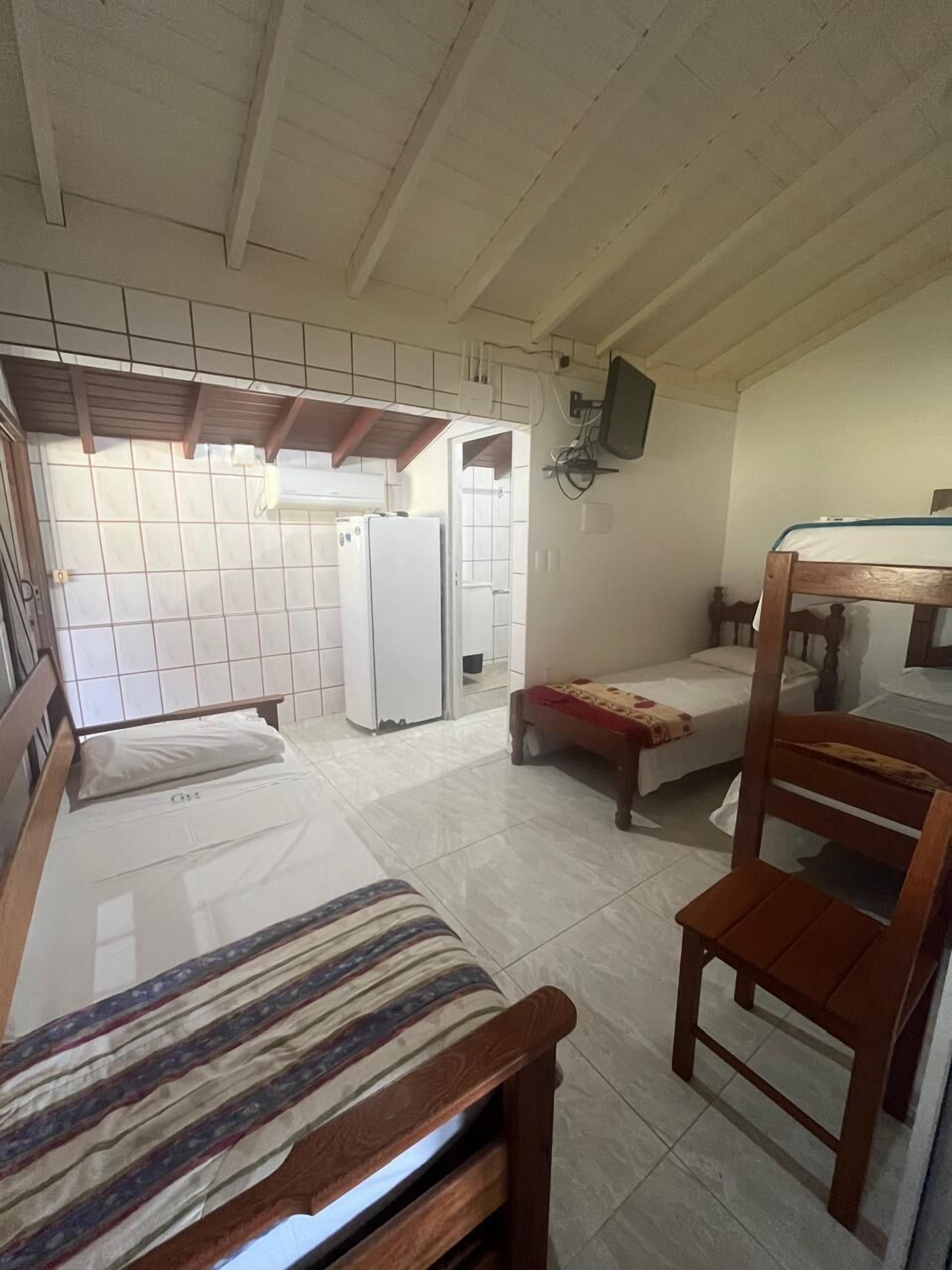 A room with two beds, a wooden chair, a television mounted on the wall, and a kitchen area with a refrigerator.
