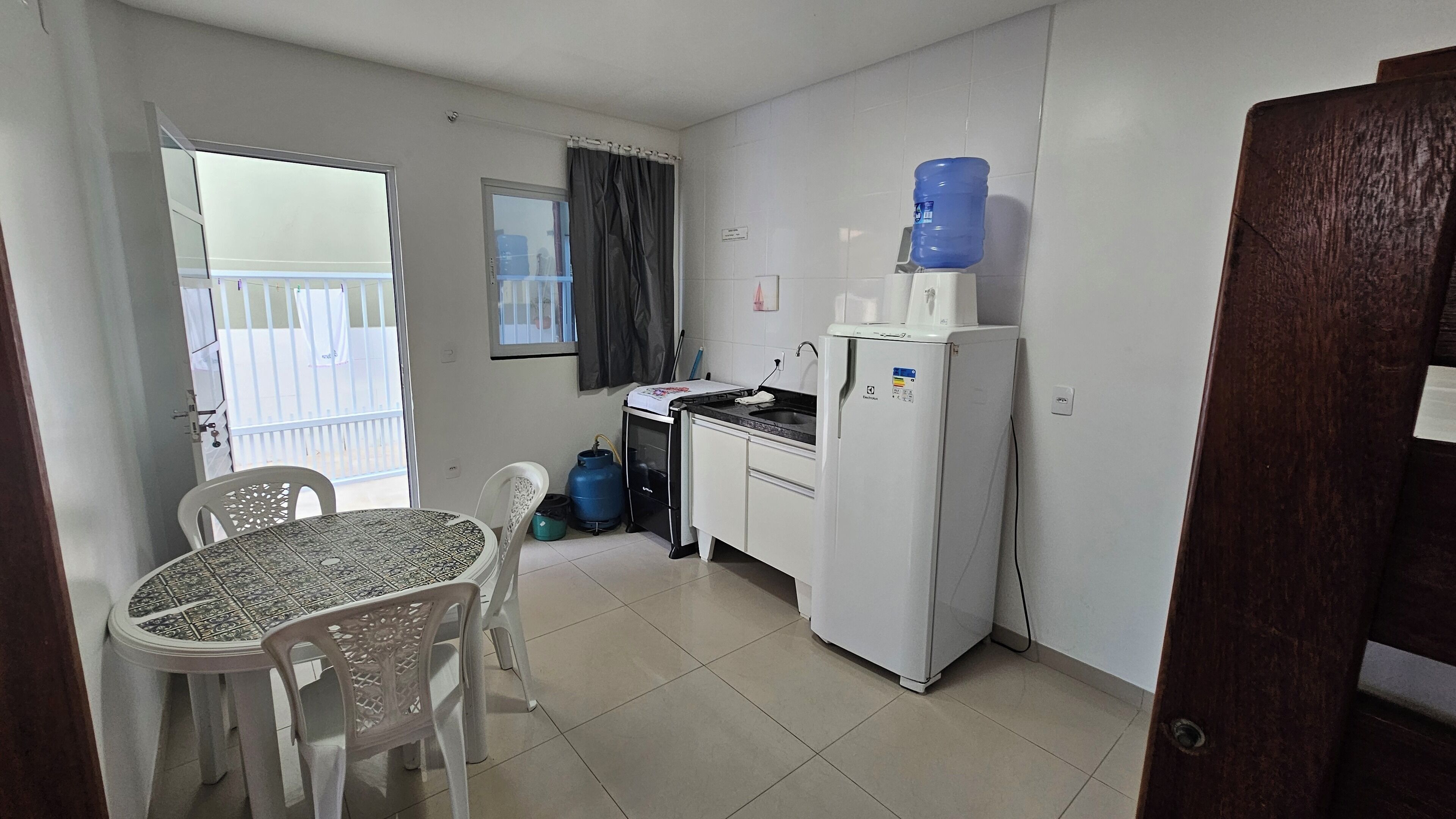 A compact kitchen with a refrigerator, sink, and a water dispenser. There's a dining table with chairs, a window with curtains, and a view of a balcony.