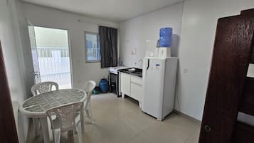 A compact kitchen with a refrigerator, sink, and a water dispenser. There's a dining table with chairs, a window with curtains, and a view of a balcony.